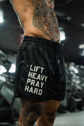 A muscular tattooed man stands in the gym, sporting Tango Charlie Apparel’s Men's Mesh Lifestyle Shorts | 5.5"- Lift Heavy Pray Hard Black, with bold slogan visible. The photo focuses on his torso and shorts, emphasizing a strong fitness lifestyle.