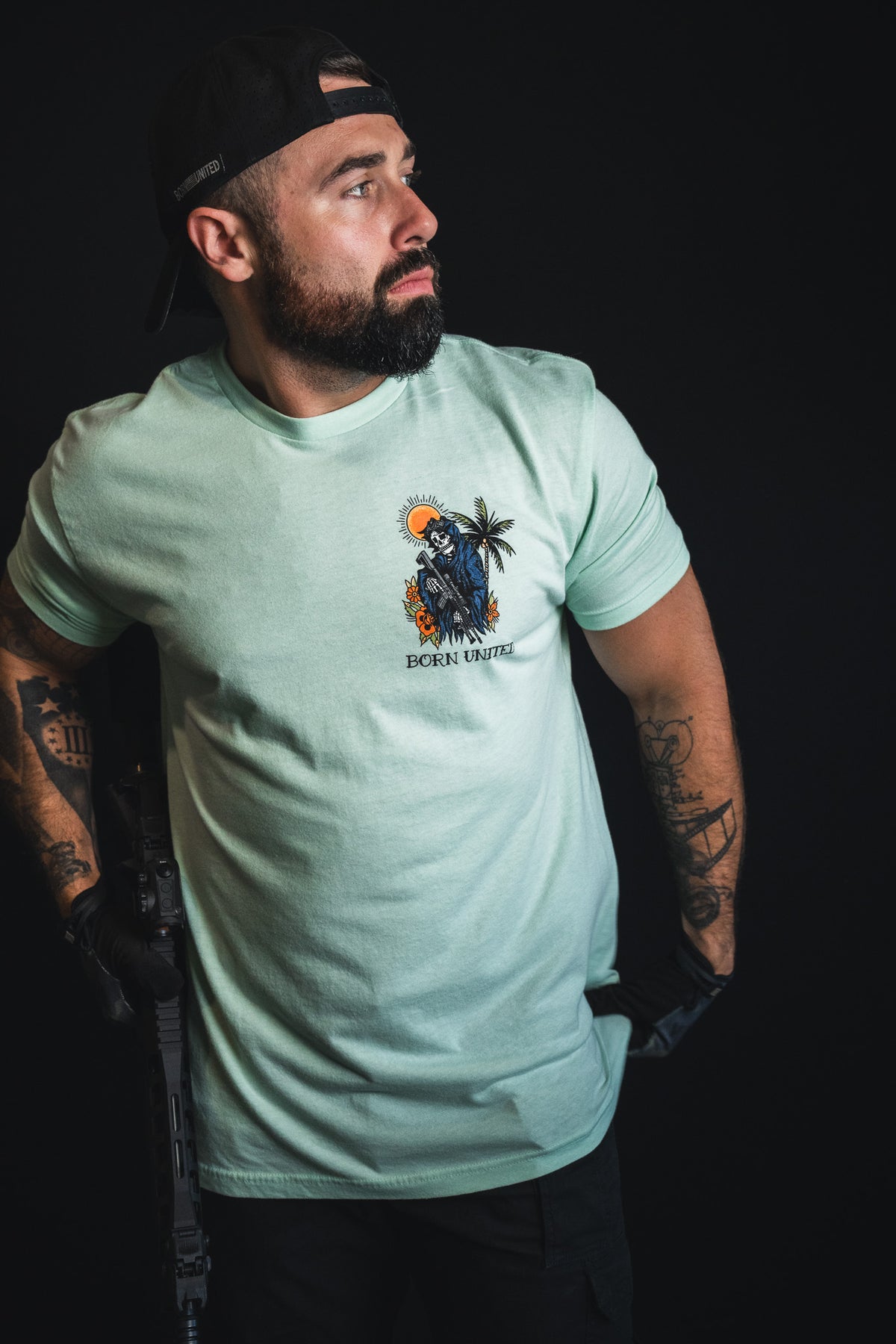 A bearded, tattooed man wearing the Born United Private Label “Always Deadly - Mint” t-shirt in an electric light green shade and a black cap looks to the side, holding a partially visible rifle against a dark background.