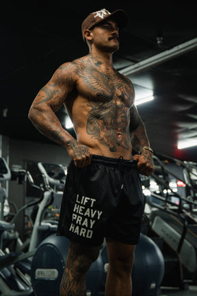 A muscular, tattooed man stands confidently in a gym wearing Tango Charlie Apparel's Men's Mesh Lifestyle Shorts | 5.5"- Lift Heavy Pray Hard Black, with exercise equipment visible in the background.