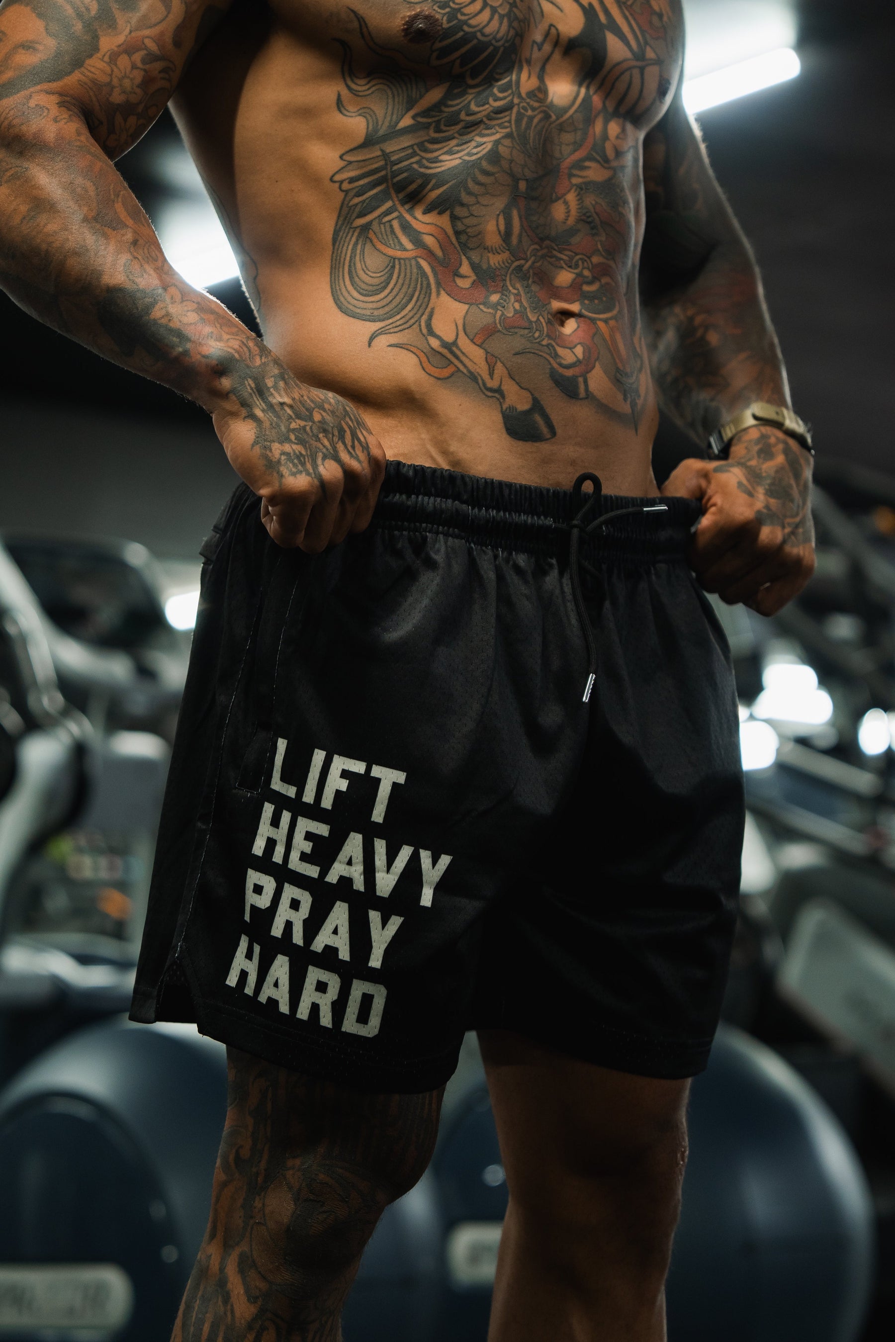 A muscular, tattooed man stands in a gym wearing Tango Charlie Apparel’s Men's Mesh Lifestyle Shorts | 5.5" - Lift Heavy Pray Hard Black, with "LIFT HEAVY PRAY HARD" boldly printed. Gym equipment is blurred in the background.