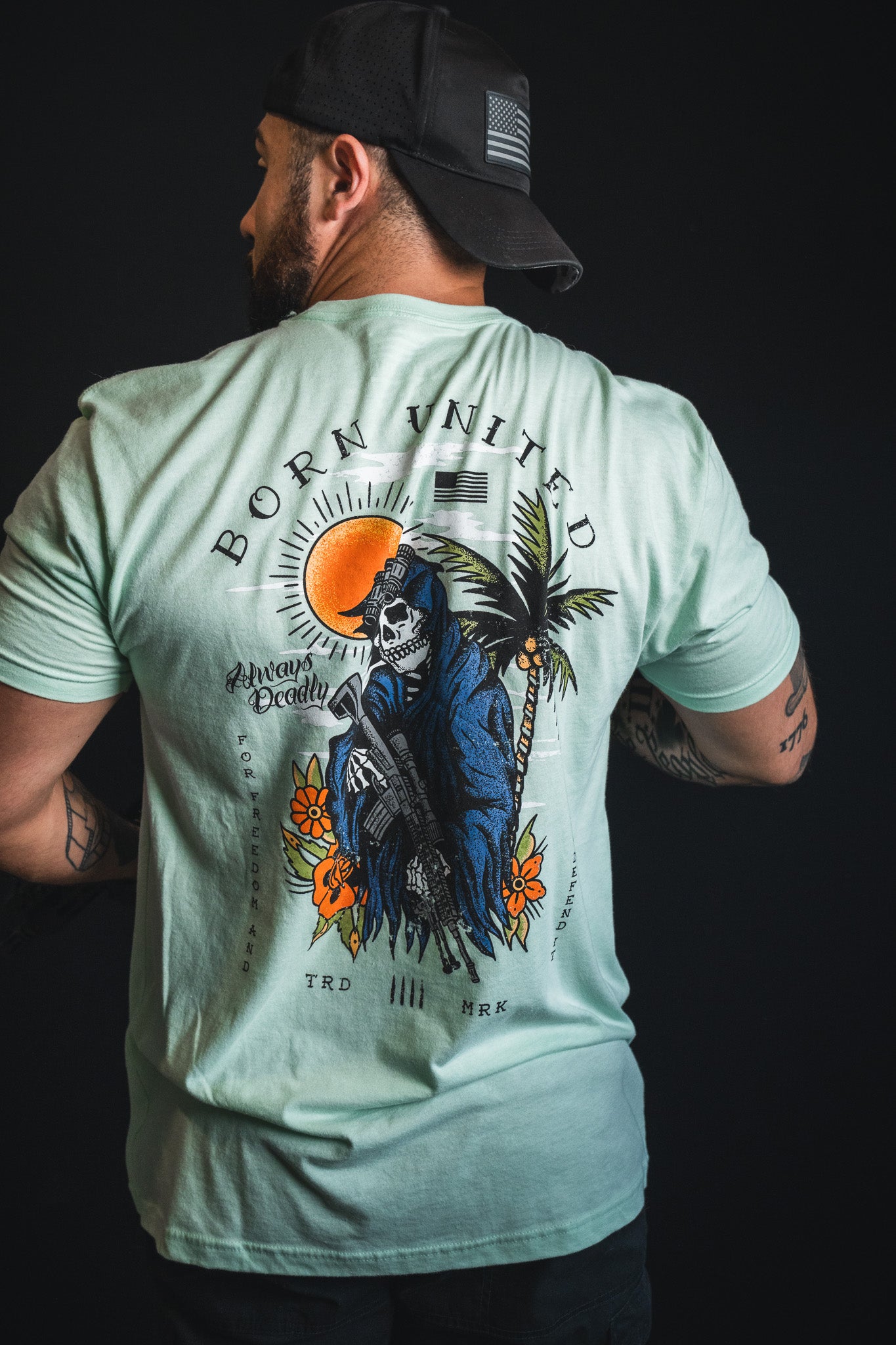 A man, seen from behind against a dark background, wears the Born United Private Label “Always Deadly - Mint” t-shirt featuring a Grim Reaper, palm trees, and “Born United” text printed on the back.