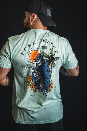 A man, seen from behind against a dark background, wears the Born United Private Label “Always Deadly - Mint” t-shirt featuring a Grim Reaper, palm trees, and “Born United” text printed on the back.