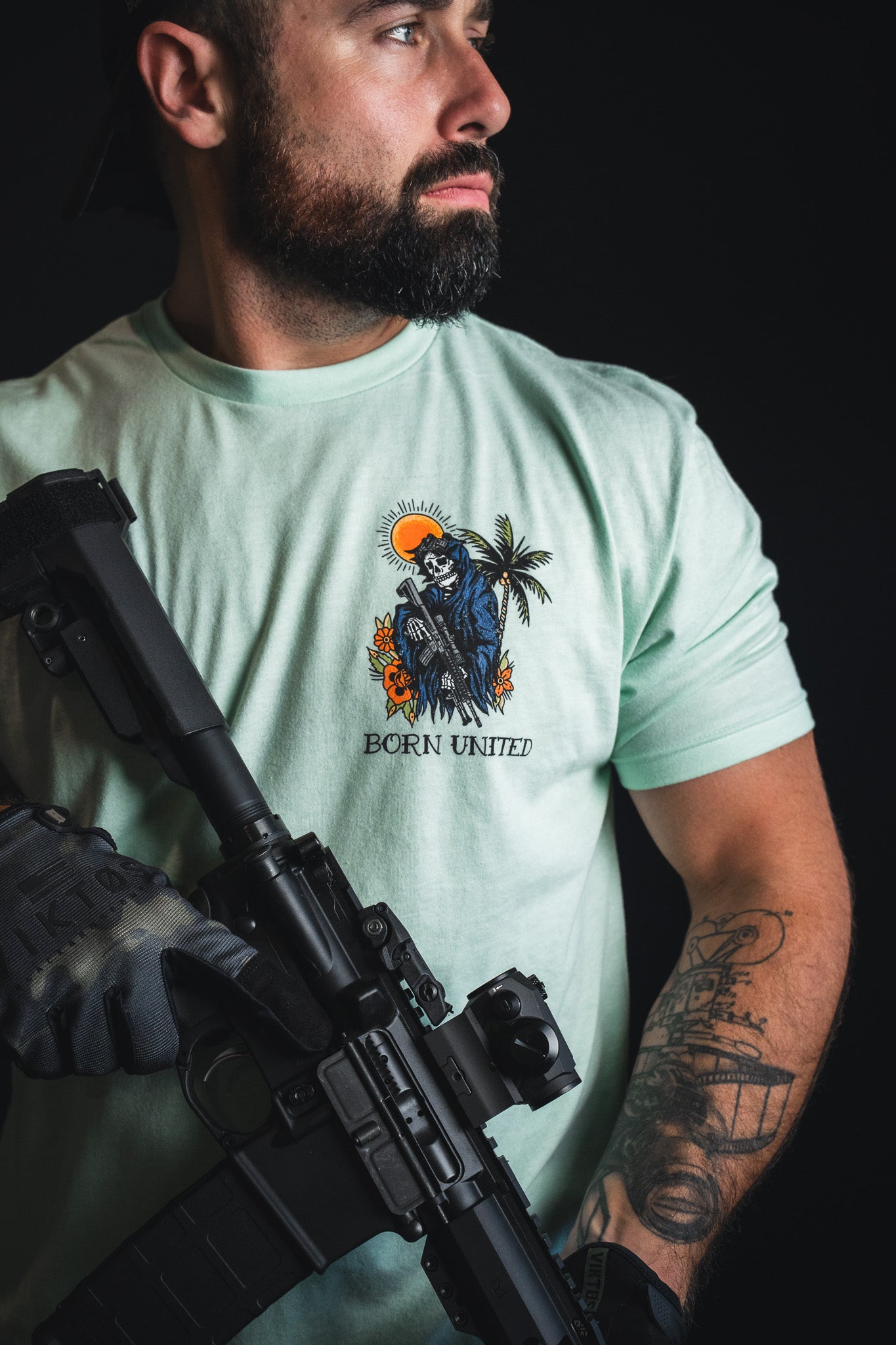 A bearded man in the “Always Deadly - Mint” t-shirt by Born United Private Label, showcasing its cool mint shade, holds a rifle and glances to the side, a forearm tattoo visible against a dark backdrop.