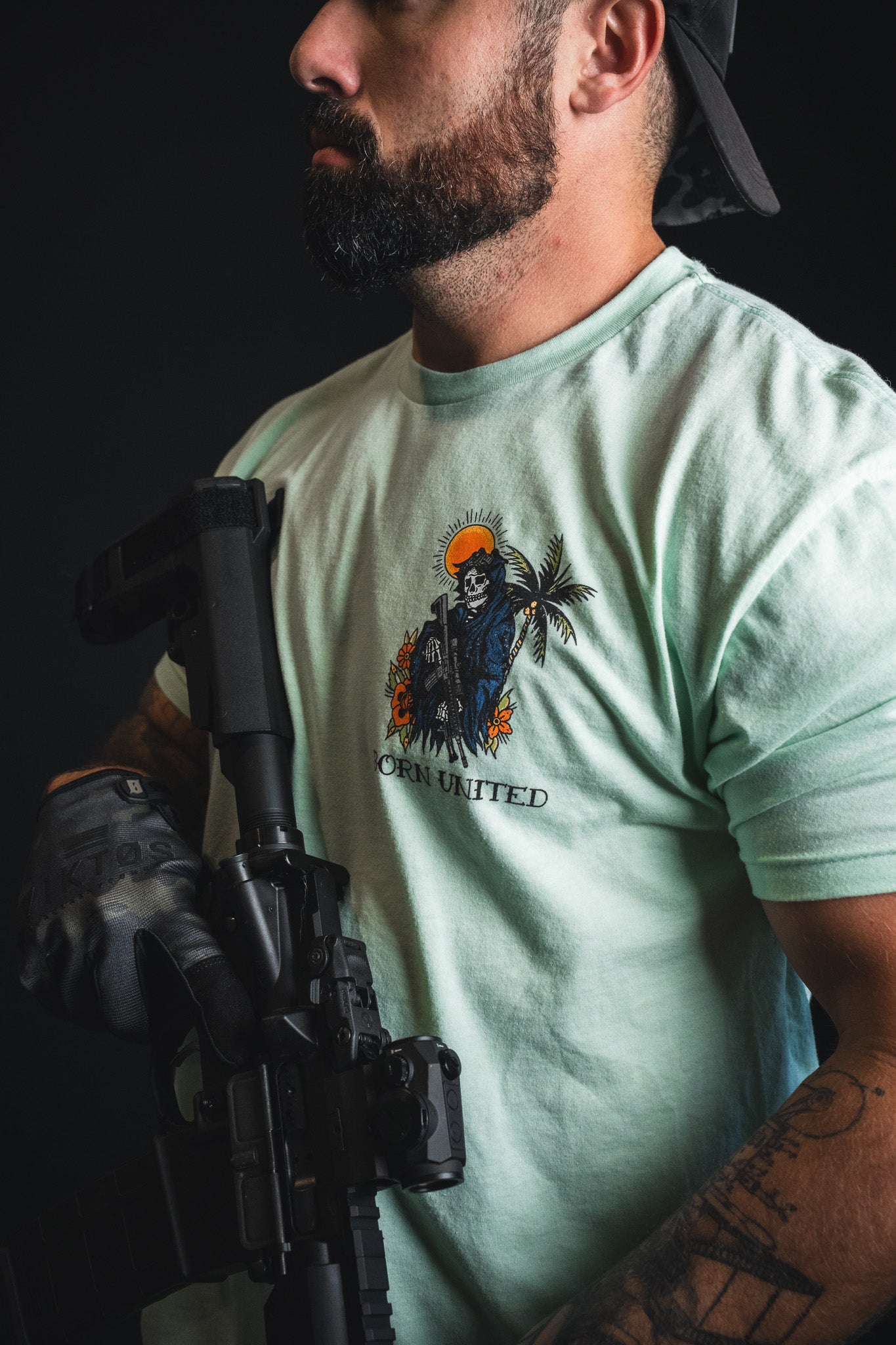 A man with a beard, wearing a backwards cap and black gloves, holds a black rifle while sporting the Born United Private Label “Always Deadly - Mint” T-shirt in an electric mint green shade with a graphic design.