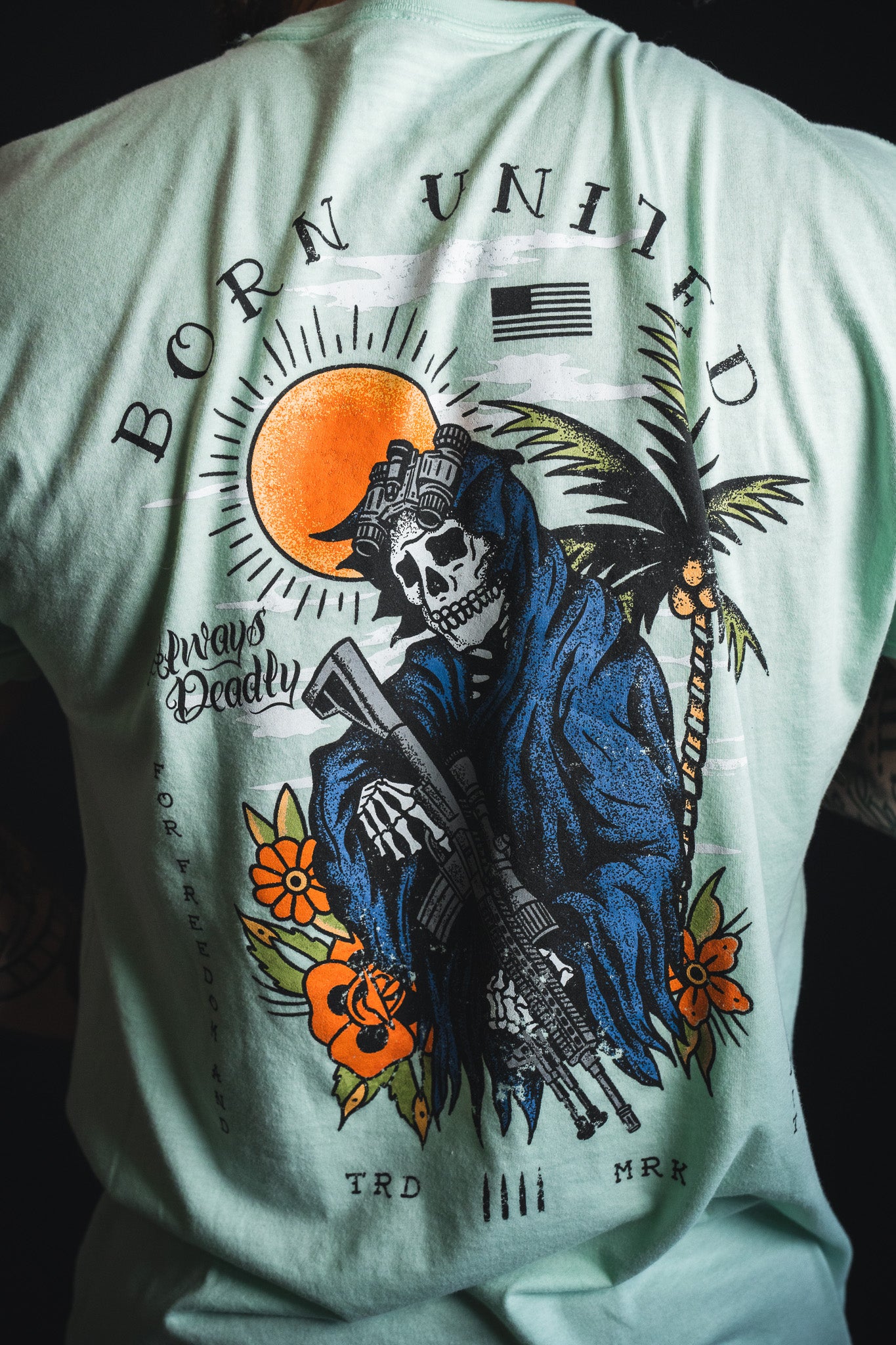 A person wearing the “Always Deadly - Mint” shirt from Born United Private Label, featuring a bold, vibrant mint color with a graphic of a skeleton in tactical gear, sun, palm trees, and the text “BORN UNITED” and “Always Steady.”.