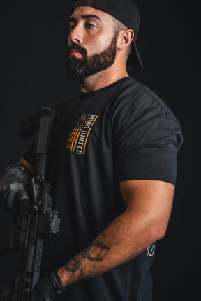 A bearded man in a black cap and gloves stands against a black background, wearing the “Walking Through Fire” premium quality t-shirt by Born United Private Label, which features a U.S. flag graphic on the chest and holds a rifle.