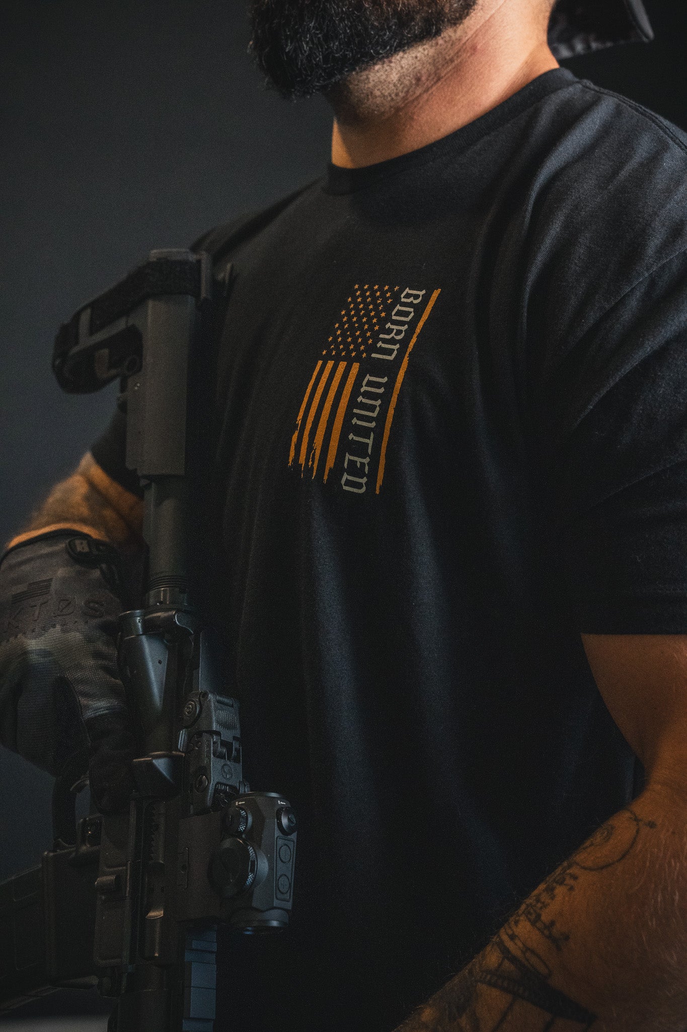 A man wearing the “Walking Through Fire” shirt by Born United Private Label, featuring an American flag graphic, holds a rifle and is partially visible from shoulders to waist.