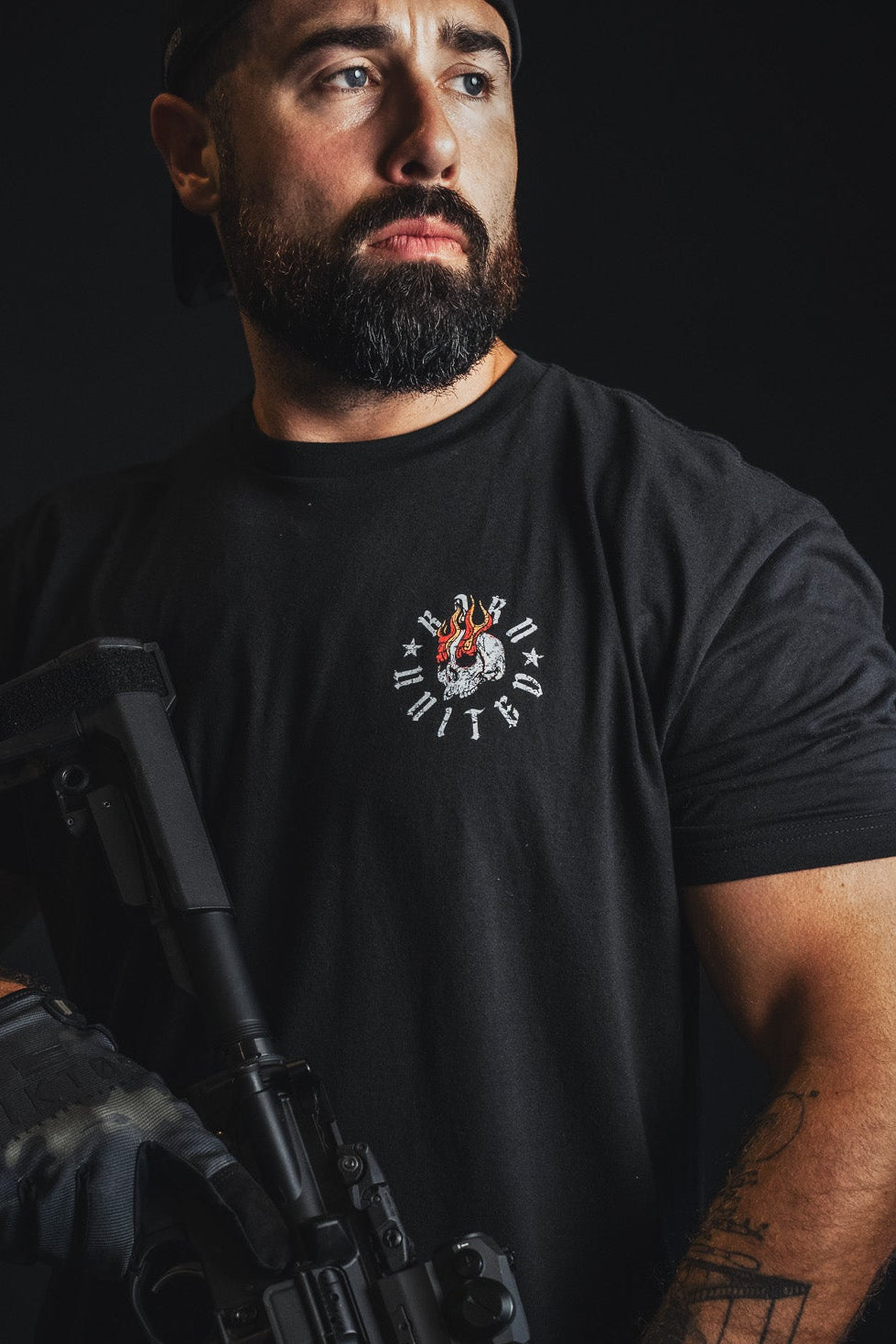 A bearded man wearing a black t-shirt with a circular 'Elite Force' logo on the chest, holding a black rifle against a dark background.