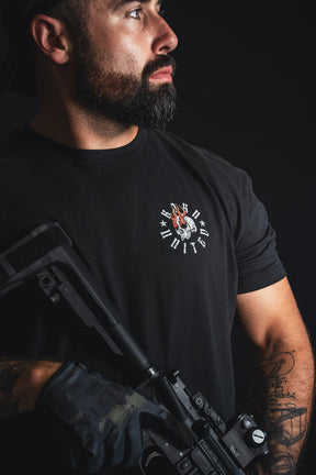 A bearded man in a black t-shirt holding a black rifle, photographed against a dark studio background. The t-shirt features a small circular graphic on the left chest with a skull and Roman numerals.