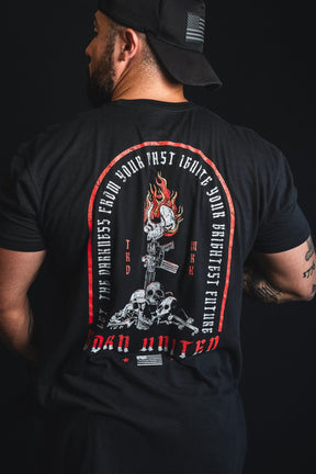 A man wearing a black t-shirt with a large back graphic, photographed from behind against a black background. The graphic features a red arched border, white text, and a detailed illustration of a skull, gun, and flames.