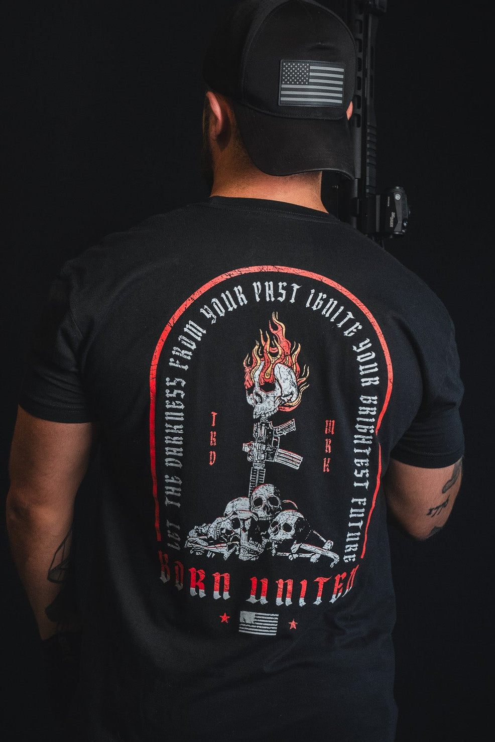 A man viewed from behind wearing a black t-shirt with a large red, white, and gray graphic print on the back, set against a solid black background.