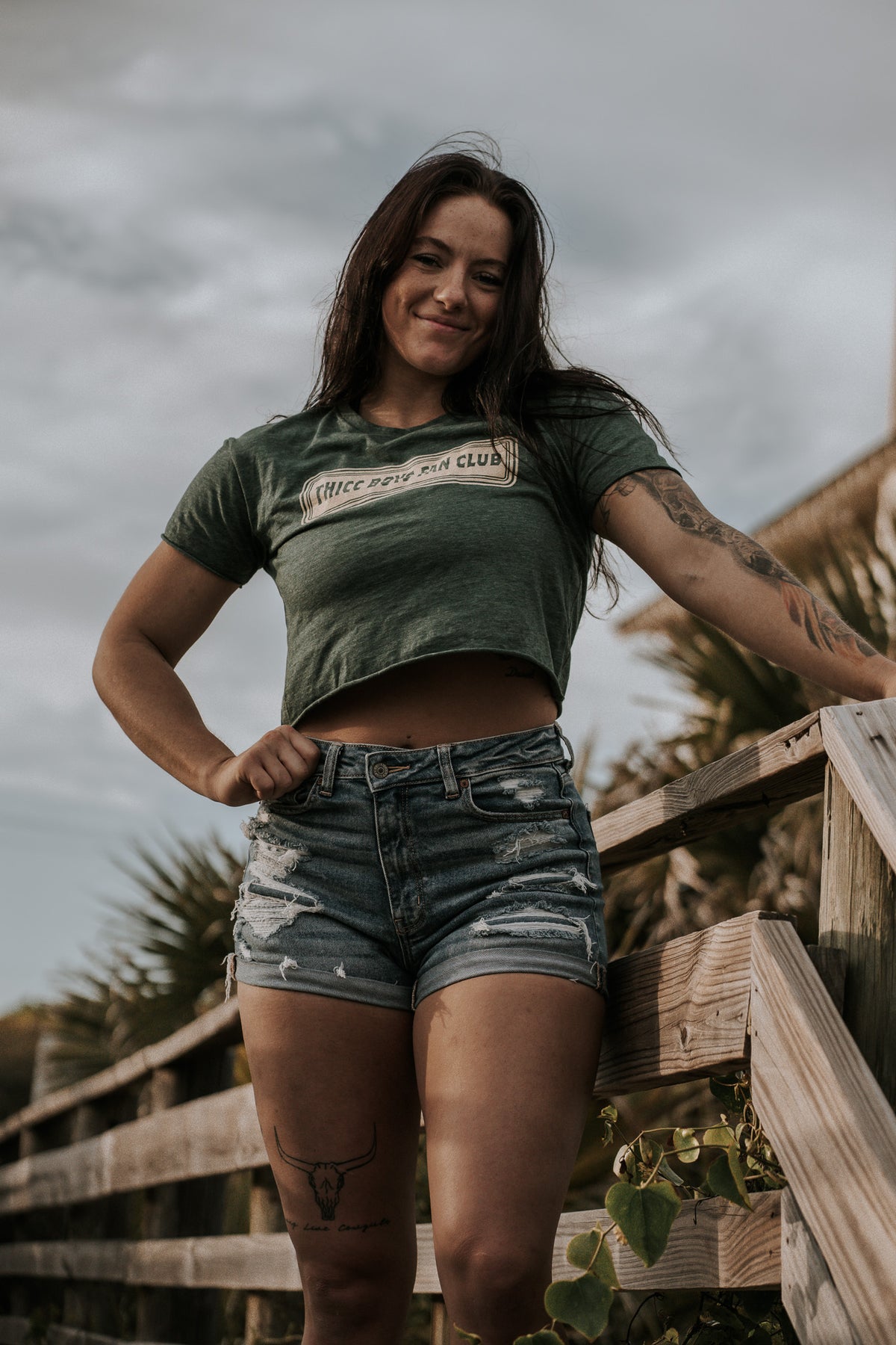 A woman with long dark hair smiles confidently on a wooden walkway, rocking the THICC Boys Fan Club Crop T-Shirt from Tango Charlie Apparel and ripped denim shorts, radiating the bold spirit of the brand by the railing.