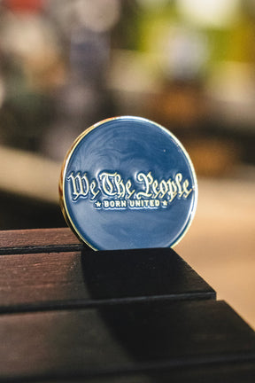 A round blue We The People Challenge Coin from the Born United Private Label collection, accented with gold trim and featuring stylized “We The People BORN UNITED” lettering, rests on a dark wooden surface.