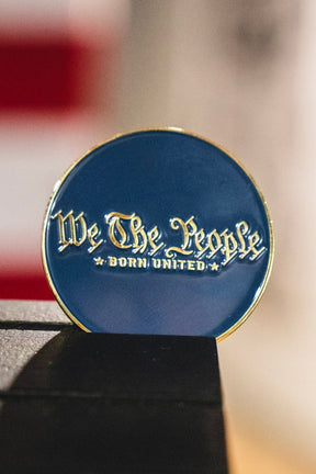 A round blue enamel pin with gold-toned lettering that reads 'We The People BORN UNITED', resting on a dark surface with a blurred background.