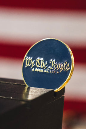 A round We The People Challenge Coin by Born United Private Label rests on a dark surface, its gold text gleaming while red and white stripes blur in the background.