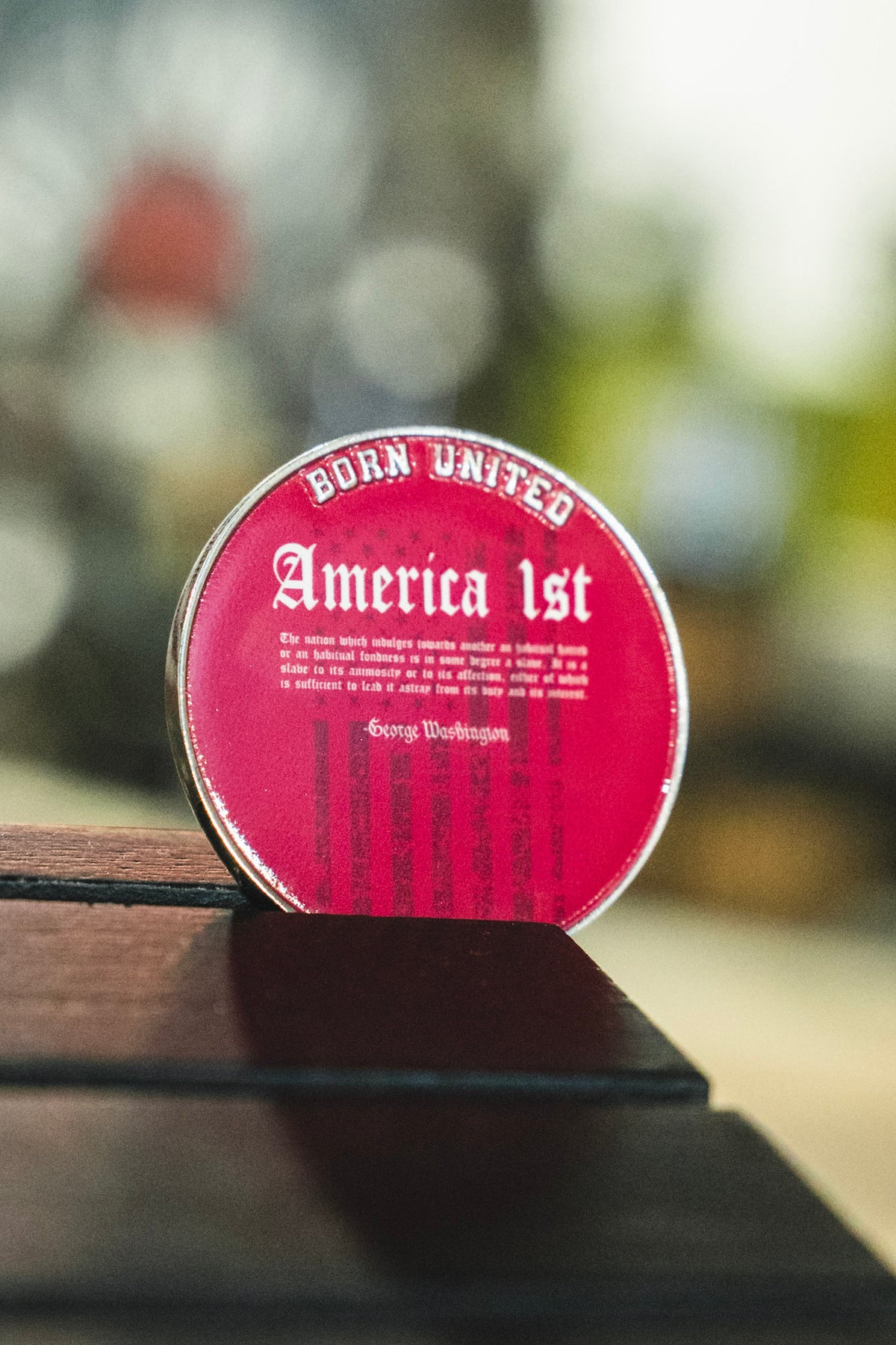 A red circular America First Challenge Coin from Born United Private Label, featuring a quote attributed to George Washington, rests against a dark wooden surface.