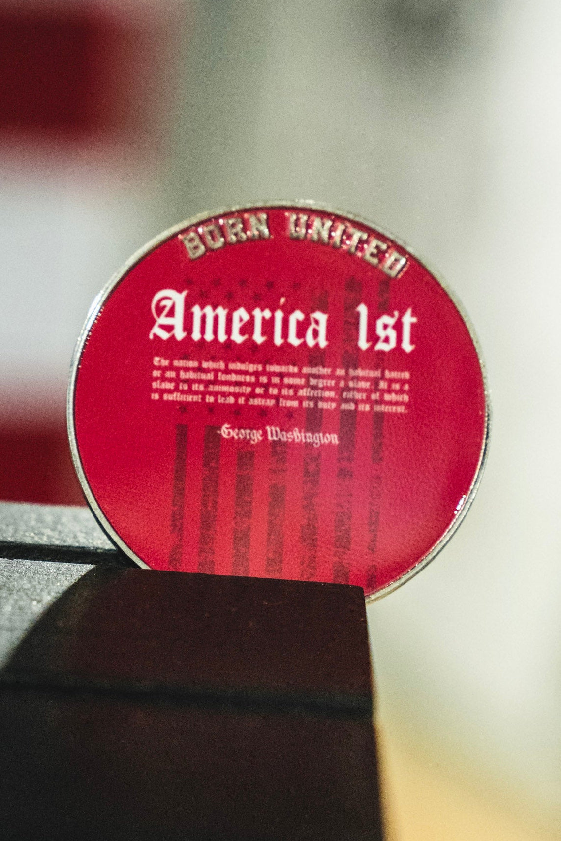 A circular red sign with white text reading 'Born United' and 'America 1st' by George Washington, photographed leaning on a dark surface with a blurred background.