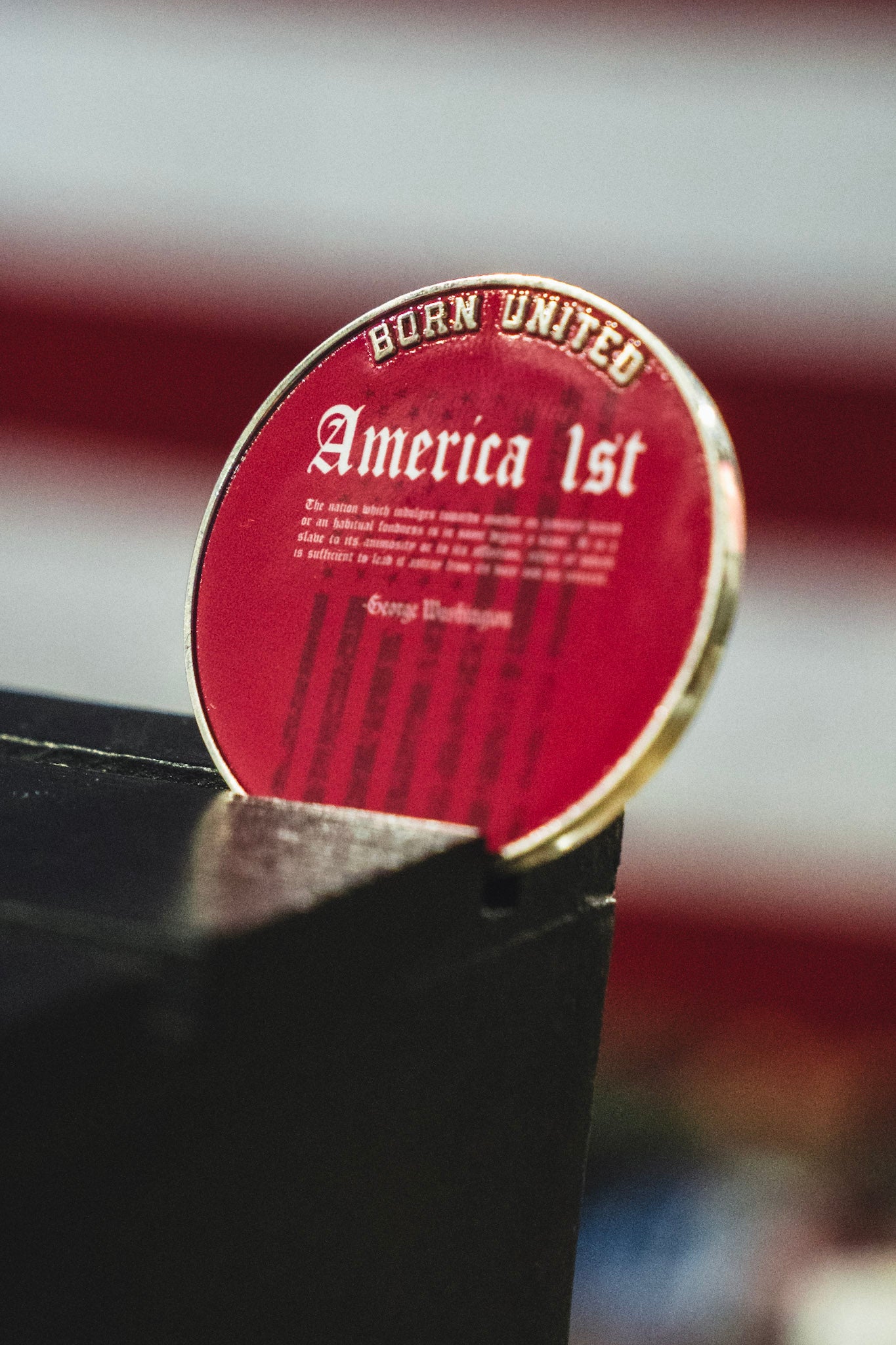 The America First Challenge Coin from Born United Private Label, featuring a round red design with “Born United” and “America First” printed on it, is propped up against a black surface with a blurred backdrop of red and white stripes.