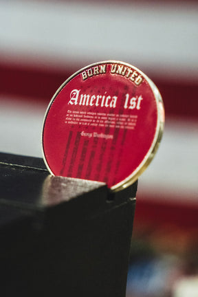 The America First Challenge Coin from Born United Private Label, featuring a round red design with “Born United” and “America First” printed on it, is propped up against a black surface with a blurred backdrop of red and white stripes.