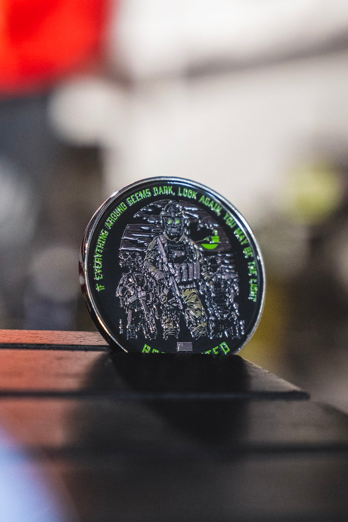 A Light in the Darkness Challenge Coin from Born United Private Label, featuring a soldier in tactical gear and green text around the edge, is displayed upright on a wooden surface.