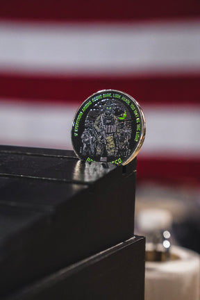 The Light in the Darkness Challenge Coin by Born United Private Label, adorned with a tactical soldier design, stands upright on a black wooden surface with an out-of-focus American flag in the background.