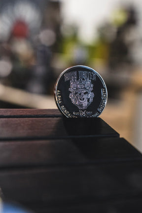 The Alpha Mentality Challenge Coin from Born United Private Label, featuring a silver finish engraved with “BORN UNITED” and an intricate mechanical design, stands upright on a dark wooden surface against a softly blurred background.