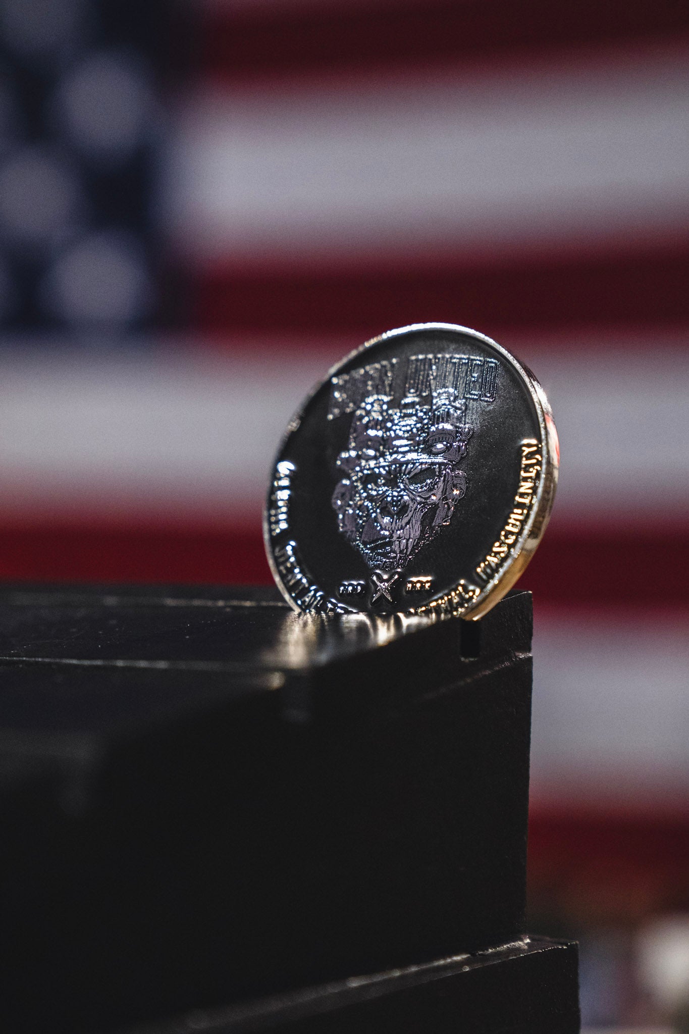 The Alpha Mentality Challenge Coin from Born United Private Label is showcased upright on a black surface, with a blurred American flag in the background, embodying pride and determination.