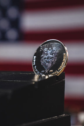 The Alpha Mentality Challenge Coin from Born United Private Label is showcased upright on a black surface, with a blurred American flag in the background, embodying pride and determination.