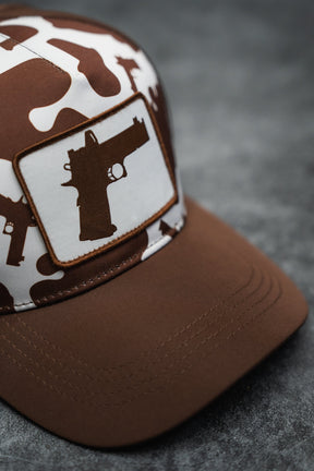 The Moo Moo Pew Hydro Snapback Hat by Tango Charlie Apparel features brown and white camouflage, a breathable design, and a handgun silhouette patch on the front, shown on a gray textured surface.