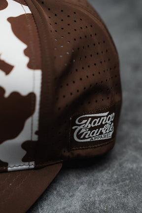 A close-up of the Moo Moo Pew Hydro Snapback Hat by Tango Charlie Apparel, featuring a brown and white camo pattern, breathable perforated panels, and a stitched side patch. The handmade cap rests on a textured gray surface.