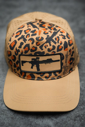 The Loaded Leopard Hydro Snapback Hat by Tango Charlie Apparel features a black and orange leopard print pattern with black rifle silhouettes on the front and brim, set against a tan base. The photo background is dark and textured.