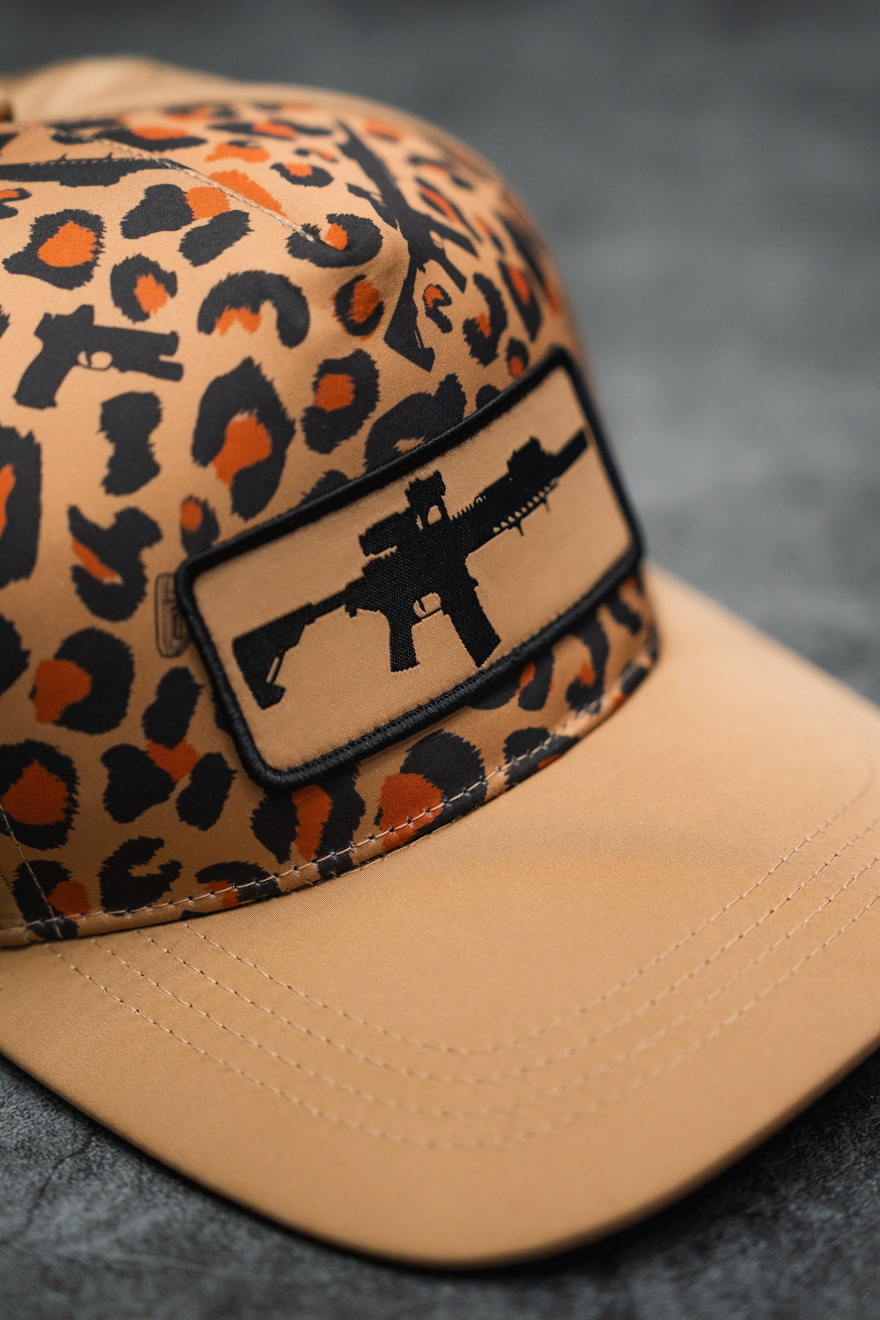 The Loaded Leopard Hydro Snapback Hat by Tango Charlie Apparel features a tan and brown leopard print with black gun silhouettes and a front patch displaying a black rifle silhouette, shown resting on a dark, textured surface.
