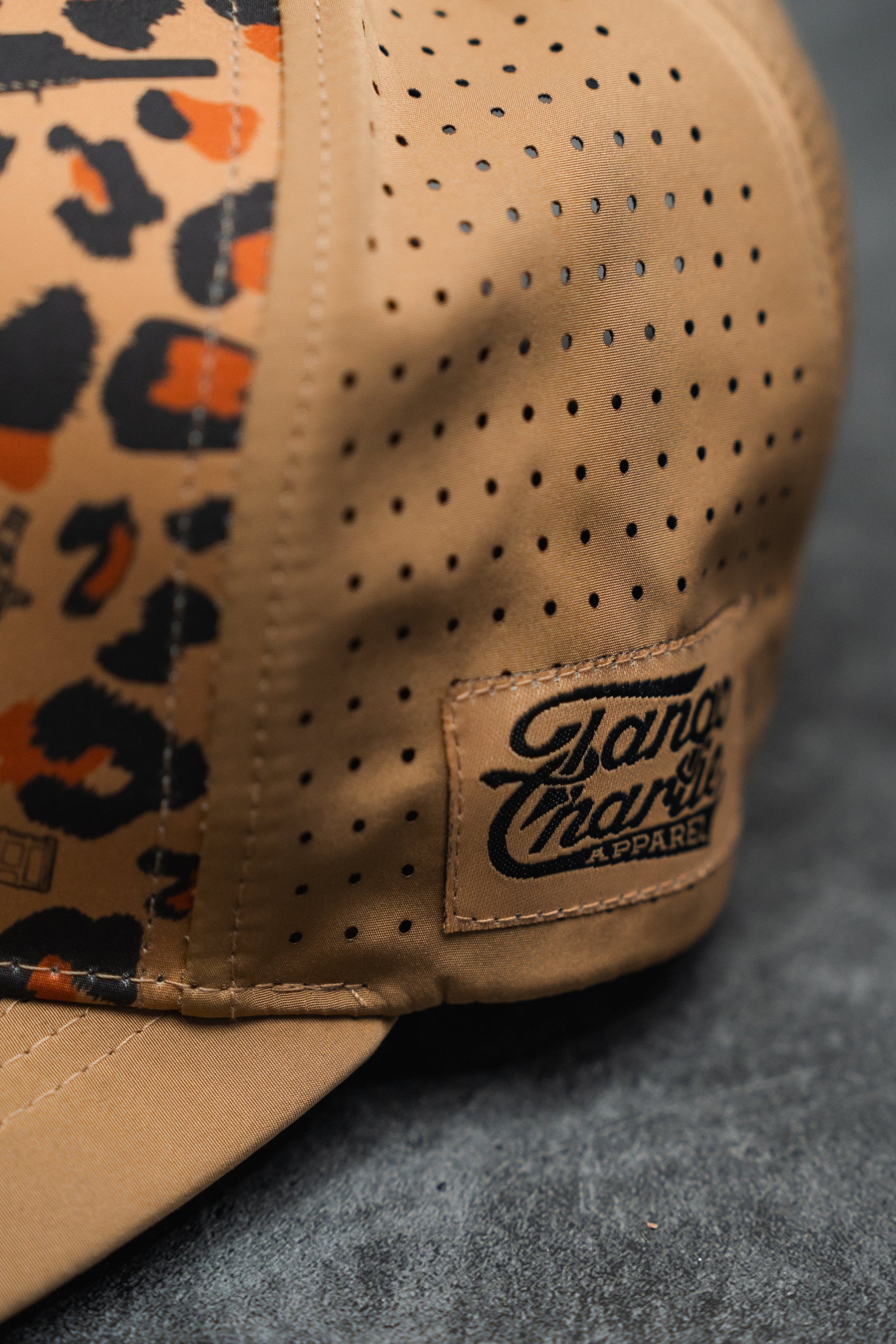 Close-up of the Loaded Leopard Hydro Snapback Hat by Tango Charlie Apparel, featuring a brown design with black and orange leopard print on one panel, ventilation holes, and a stitched label. The cap rests on a dark textured surface.