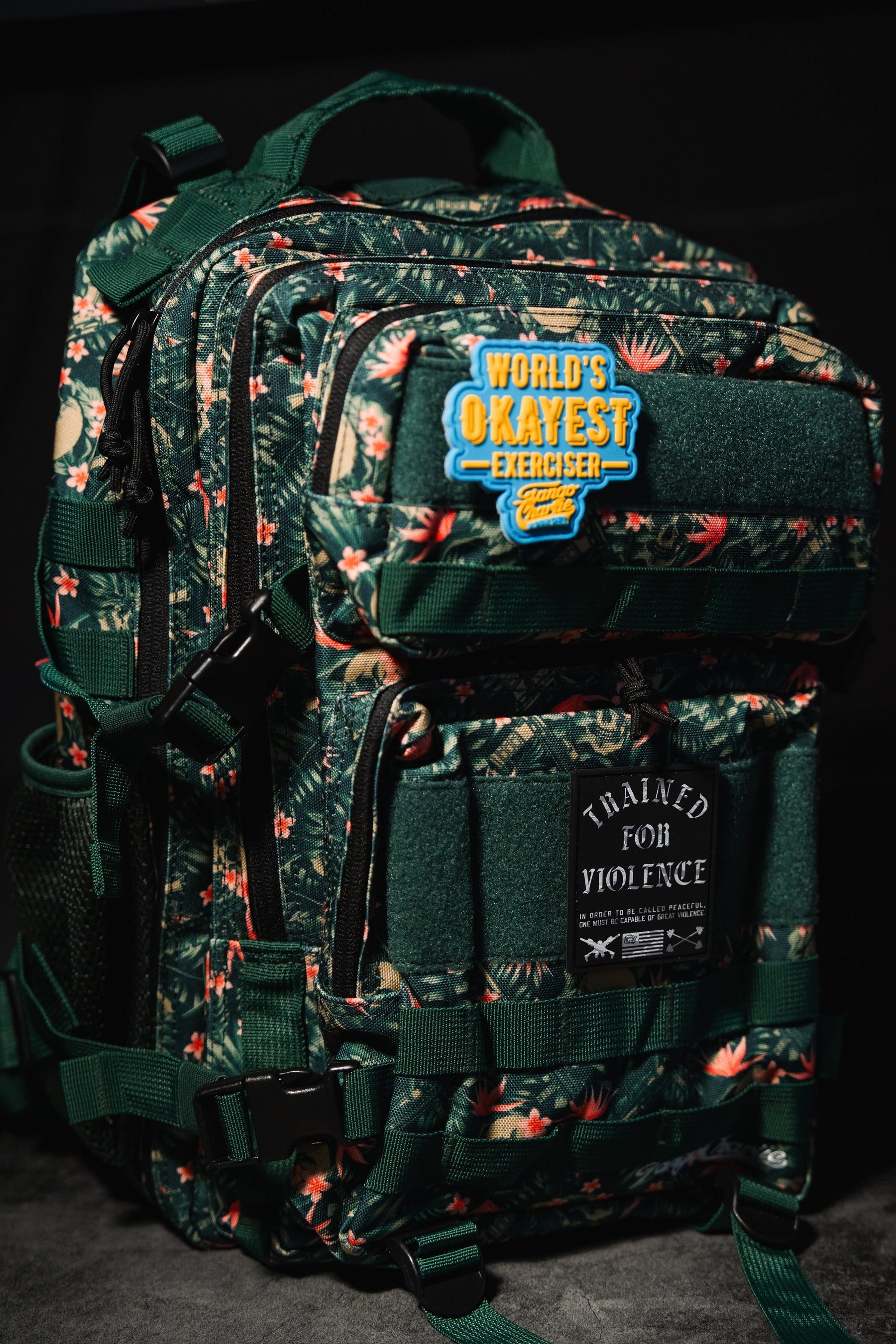 Aloha AR - Tac Pac 25L by Tango Charlie Apparel: a dark green floral-patterned hunting backpack with two patches—WORLDS OKAYEST EXERCISER and TRAINED FOR VIOLENCE—plus multiple zippers and straps.