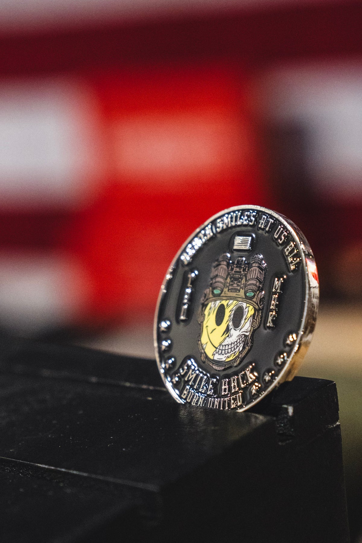 A close-up of the Smile Back Challenge Coin by Born United Private Label, showcasing a skull adorned with gears and wires subtly grinning, alongside the text “MIKE BUCK BORN UNITED,” set against a blurred red and white background.