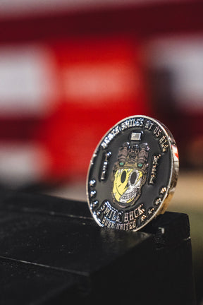 A close-up of the Smile Back Challenge Coin by Born United Private Label, showcasing a skull adorned with gears and wires subtly grinning, alongside the text “MIKE BUCK BORN UNITED,” set against a blurred red and white background.