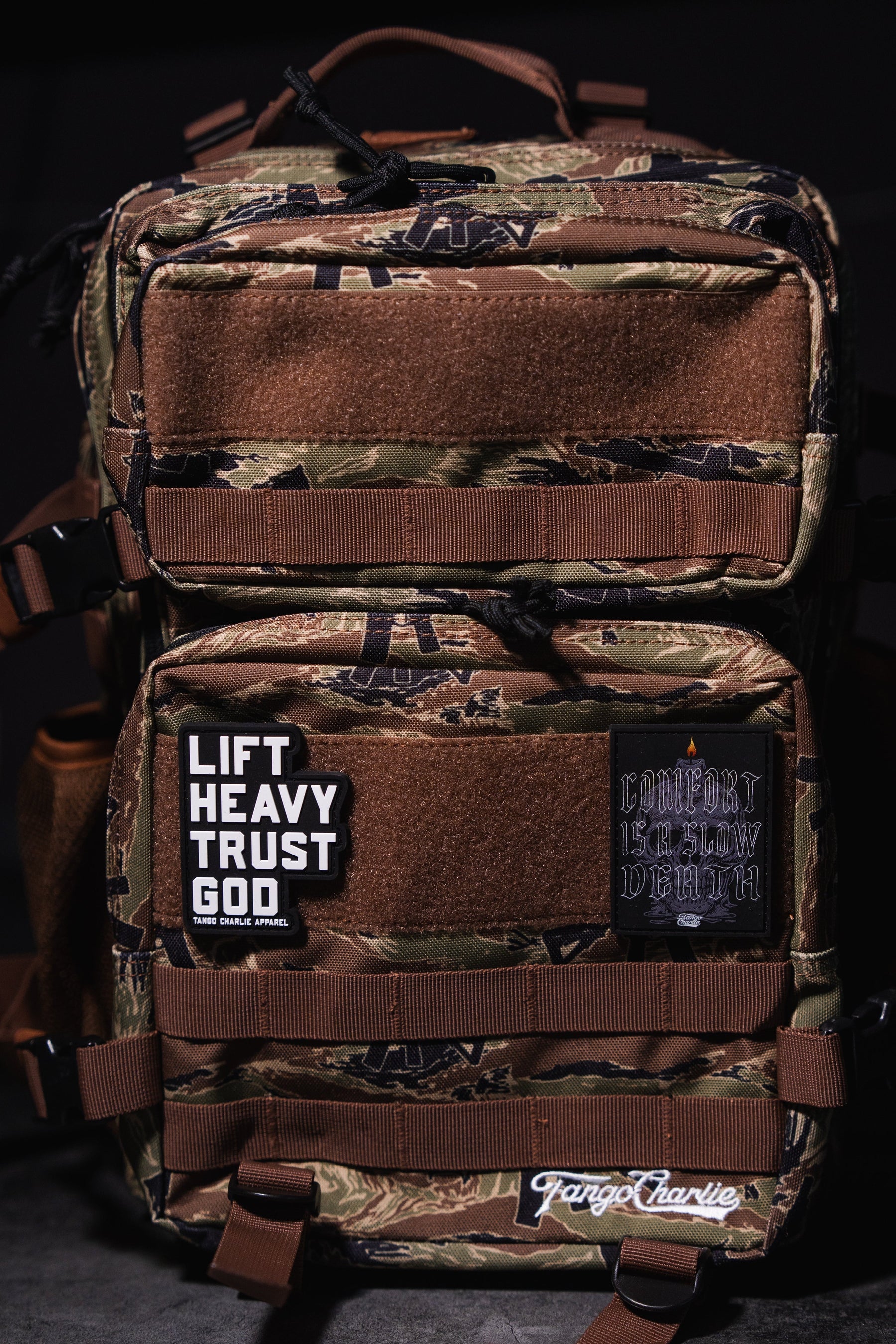 The Snipes & Stripes - Tac Pac 25L by Tango Charlie Apparel is a tactical backpack with brown straps, a camouflage design, and bold patches: "LIFT HEAVY TRUST GOD" in white and "COMFORT IS SLOW DEATH" in gothic text.