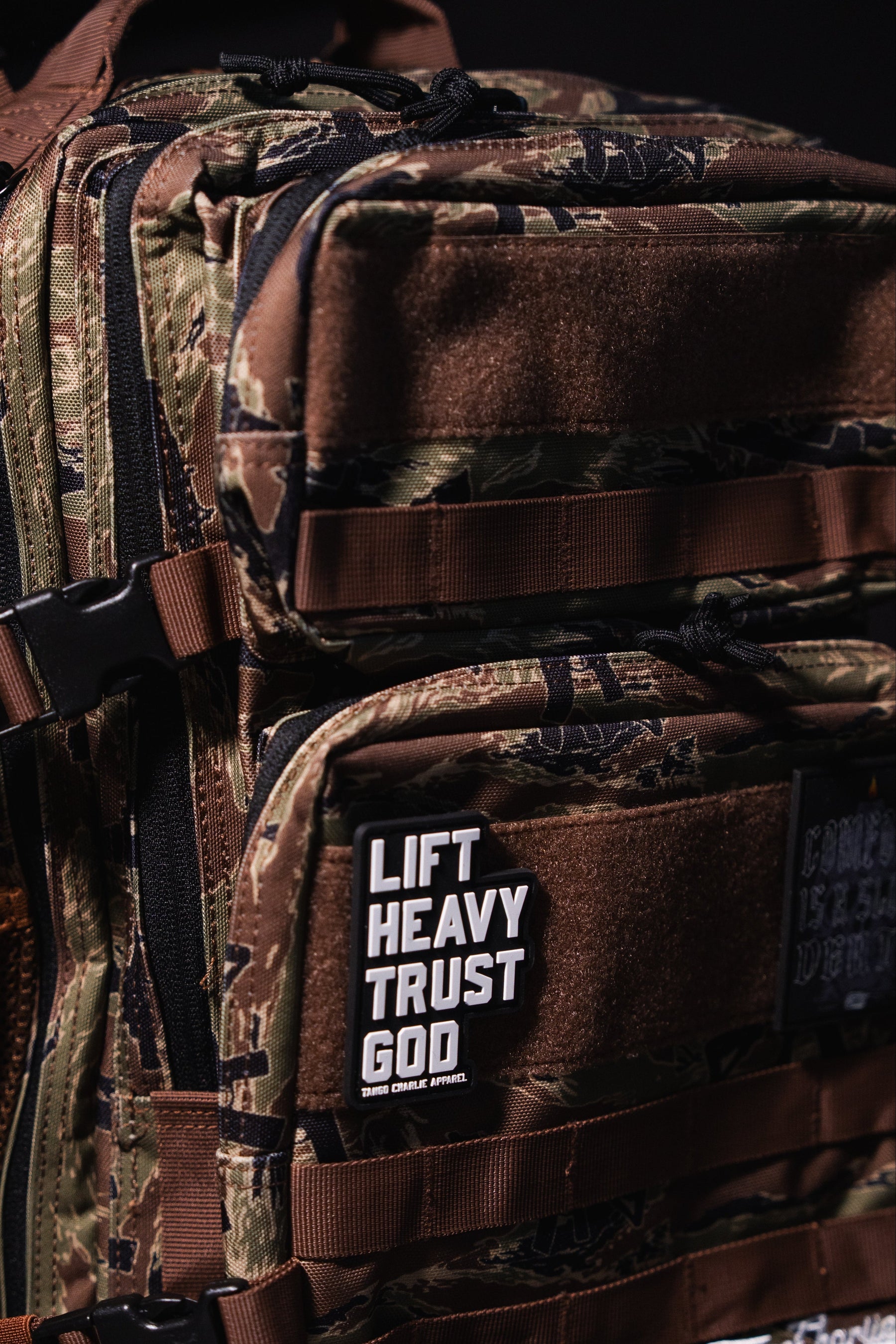 A close-up of the Snipes & Stripes - Tac Pac 25L by Tango Charlie Apparel shows a camouflage tactical backpack with brown straps. A front patch reads “LIFT HEAVY TRUST GOD” in bold white letters. The dark background highlights its rugged style.