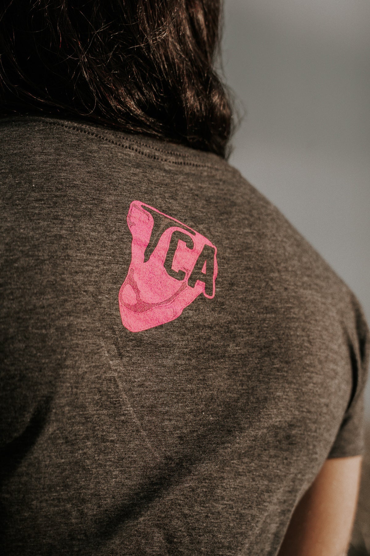 A close-up shows the back of a person wearing a charcoal THICC Boys Fan Club Crop T-Shirt by Tango Charlie Apparel. The shirt features a pink graphic with TCA lettering, partially covered by the persons dark hair.