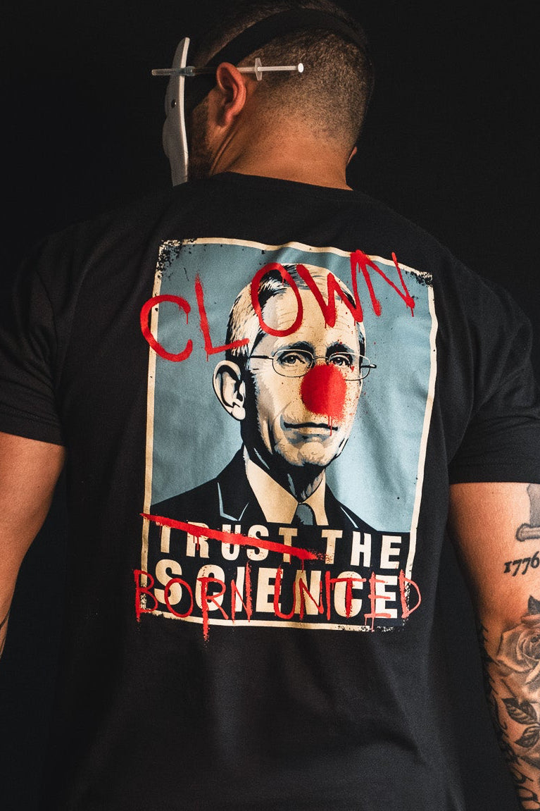 A man stands against a black background wearing Born United Private Label’s “The Fraud Show” shirt, which features a graphic of a suited man with red clown features and text reading “CLOWN” and “TRUST THE SCIENCE” crossed out—suggesting skepticism towards trusting science.