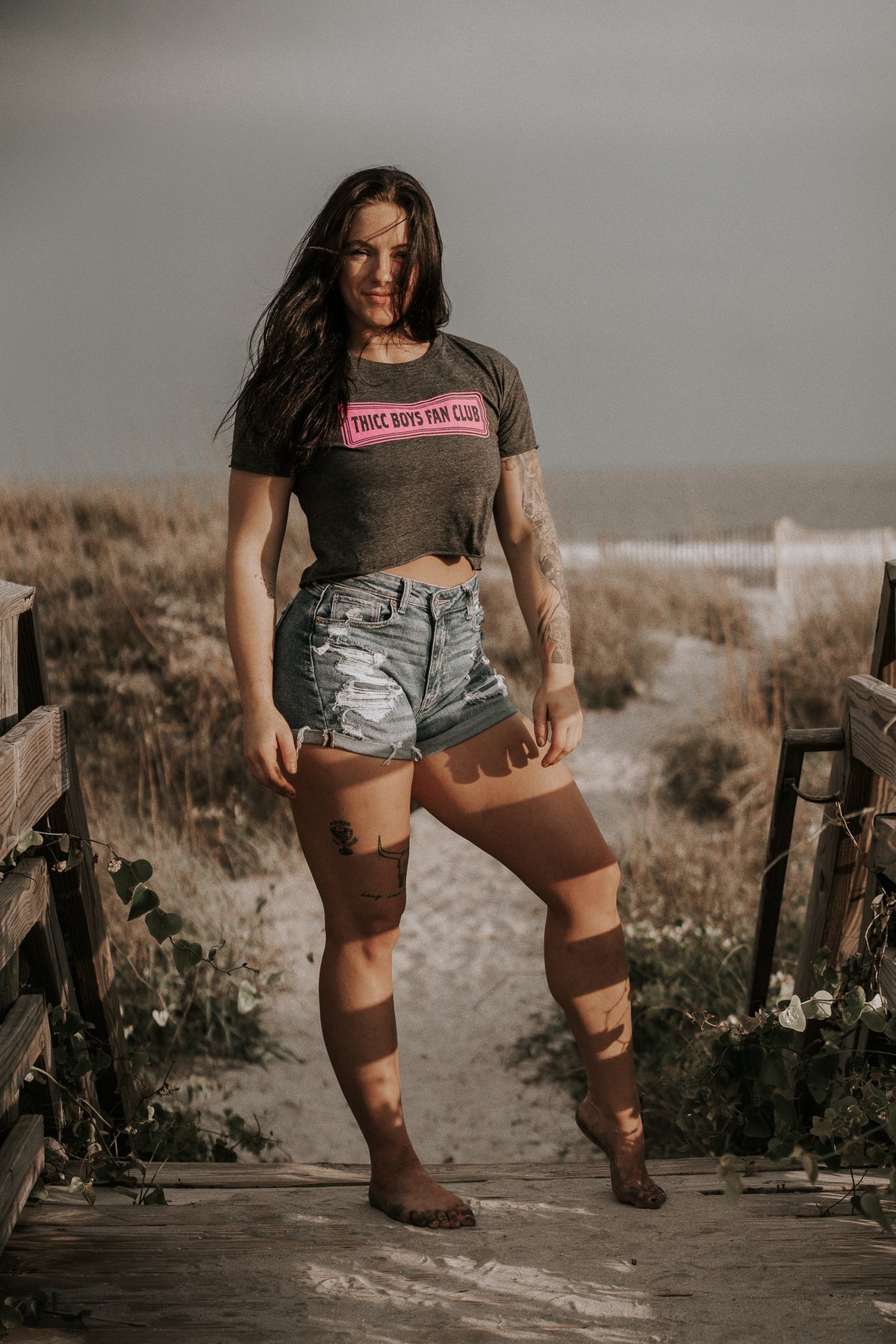 A woman with long dark hair stands barefoot on a sandy beach path, wearing the Tango Charlie Apparel THICC Boys Fan Club Crop T-Shirt in Charcoal and ripped denim shorts, with grassy dunes and the ocean behind her.