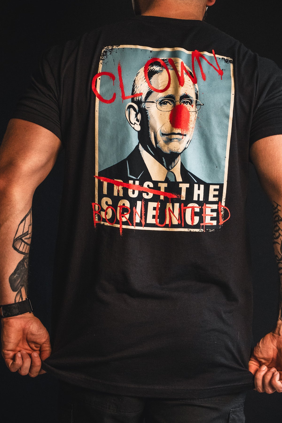 A person wearing Born United Private Label’s “The Fraud Show” shirt, featuring a doctored image of a man with a red nose, bold red graffiti text saying “CLOWN,” and modified lettering reading “TRUST THE FORBIDDEN”—a satirical nod to trusting science while some plead the 5th.