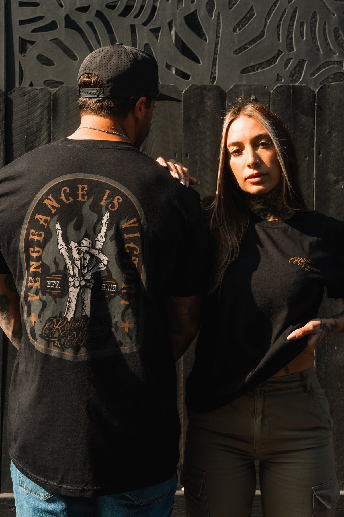 A man and woman stand before a textured black wall. The man wears a Vengeance Tee from Relentless Betrayal, symbolizing resilience, with a cap on. The woman, in her black tee and khaki pants, gazes confidently at the camera while gripping her shirt.