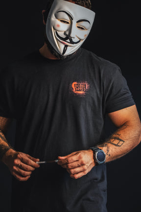 Wearing a Guy Fawkes mask and The Fraud Show T-shirt by Born United Private Label, a person holds a syringe against a dark backdrop, prompting viewers to reflect on where their trust truly lies—in science, society, or secrecy.