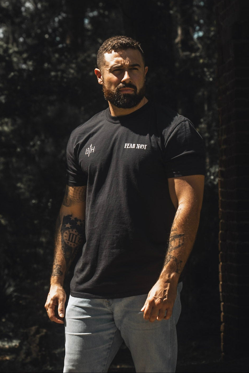 A tattooed, bearded man stands outdoors among trees and shadows, wearing the 13th Fear Not Premium Tee in black with light jeans—a shirt drawing spiritual inspiration from Isaiah 41:10.