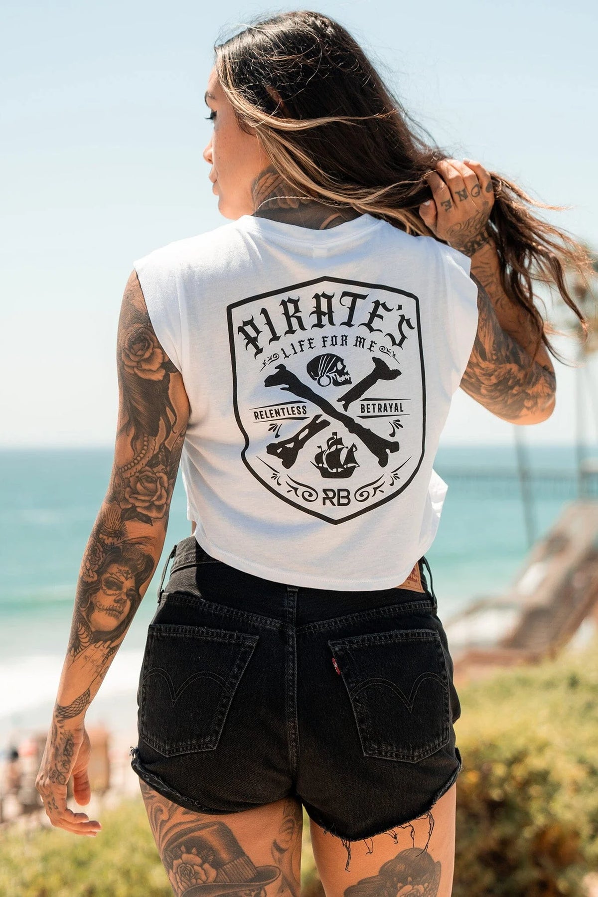 A tattooed woman with long hair stands at the beach, facing away, embodying freedom in her sleeveless Relentless Betrayals Pirates Life Womens White Crop featuring a pirate ship illustration, paired with black denim shorts. The ocean and sky stretch endlessly behind her.