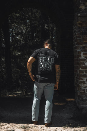 A person stands outdoors facing away from the camera, wearing the 13th brand Fear Not Premium Tee—a black T-shirt featuring white text inspired by Isaiah 41:10—paired with light blue jeans and brown shoes, near a brick structure and trees. The scene captures a quiet moment of faith and spiritual inspiration.