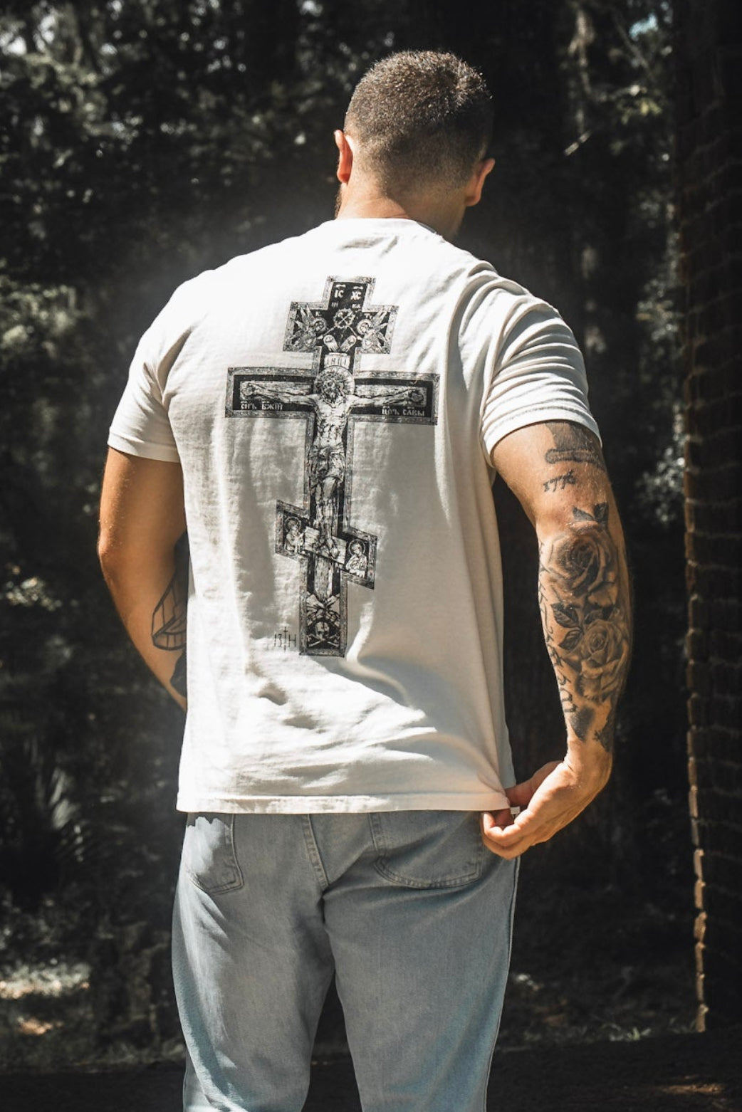 A man stands outdoors with his back to the camera, wearing the 13th Paid in Full Premium Tee, which features a large ornate cross design that beautifully symbolizes faith and redemption, paired with light blue jeans.