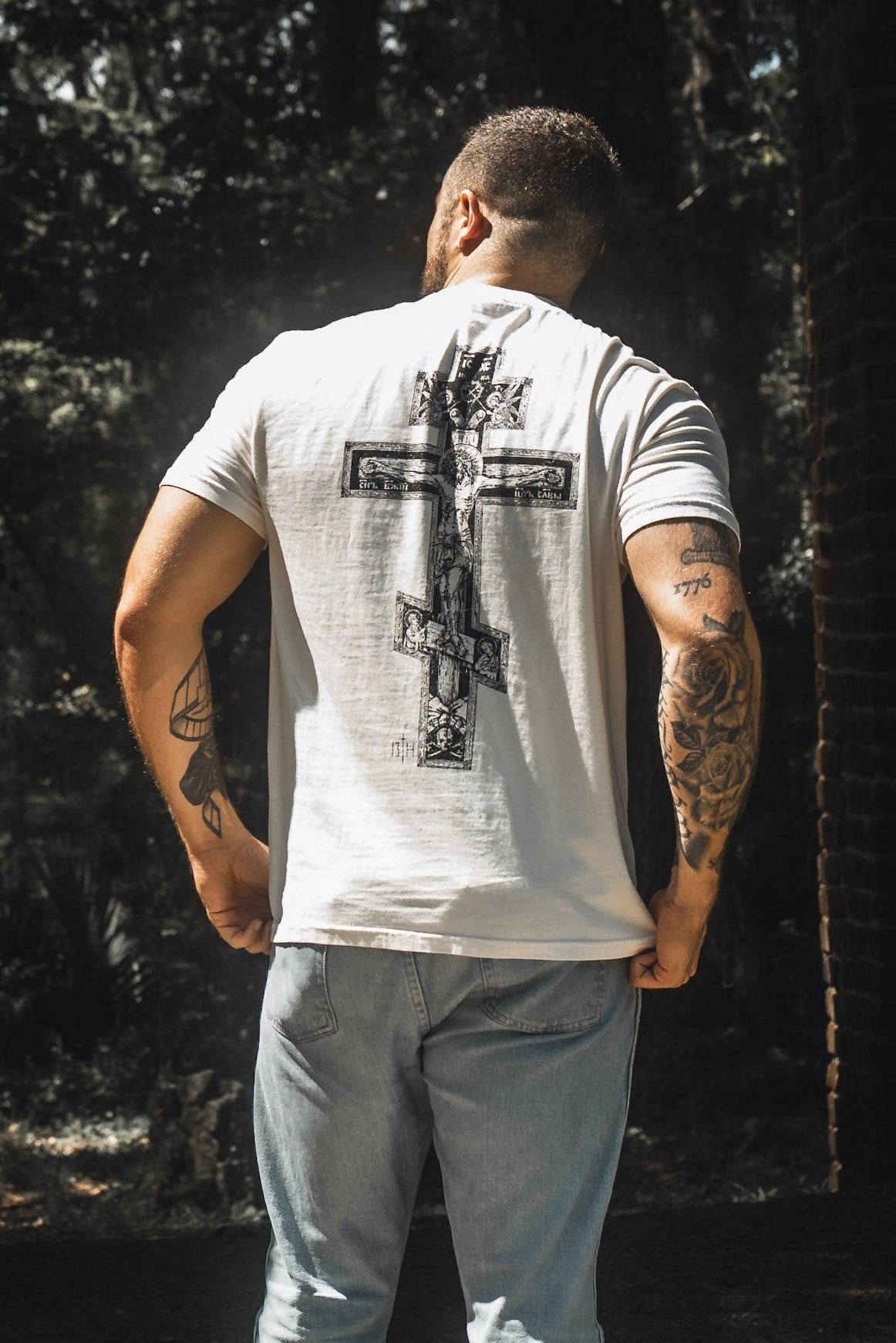 A person stands outdoors in the shade, facing away, wearing the 13th Paid in Full Premium Tee featuring a bold graphic of Jesus on the cross across the back, paired with light blue jeans. Their tattooed arms and choice of apparel create a powerful statement of faith and redemption.