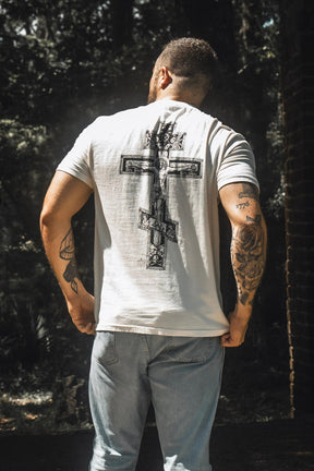 A person stands outdoors in the shade, facing away, wearing the 13th Paid in Full Premium Tee featuring a bold graphic of Jesus on the cross across the back, paired with light blue jeans. Their tattooed arms and choice of apparel create a powerful statement of faith and redemption.